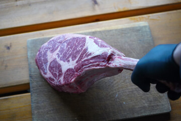 Raw t bone steak is on a wooden board on black background, next to salt, pepper and herbs 