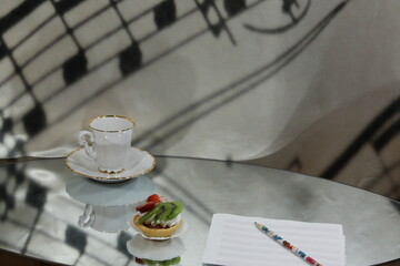 Cake with slices of strawberry kiwi berries in a tartlet on a mirrored table with a sheet of music with a pencil and a white coffee cup with a saucer.Art creative background