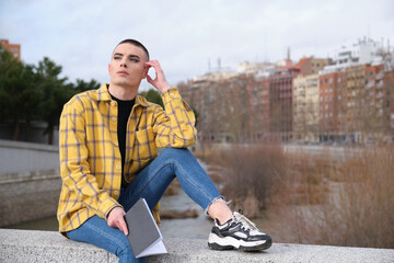 Handsome young man wearing make up, looking thoughtful with a book in his hands sitting on a wall....