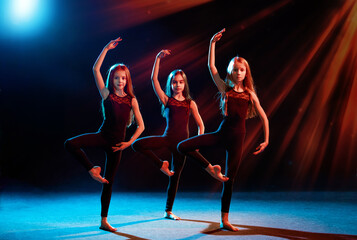 group of three ballet girls in tight-fitting costumes dance against black background with their long hair down, silhouettes illuminated by color sources.