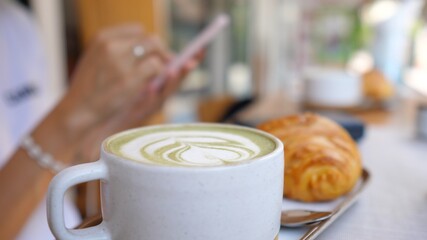 Hand taking mug with matcha green tea latte. Smartphone blurred in the background