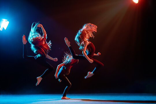 group of three ballet girls in tight-fitting costumes jumping on black background with their long hair down, silhouettes illuminated by color sources