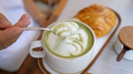 Top view of spoon taking froth off the matcha latte. Healthy dairy free drink served with fresh croissant 