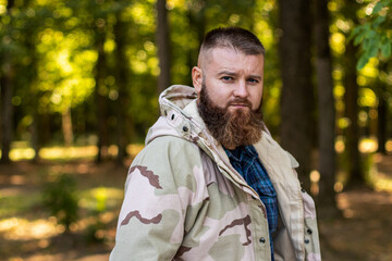 A man with a beard in a military jacket looks at the camera, in the background forest, soft focus