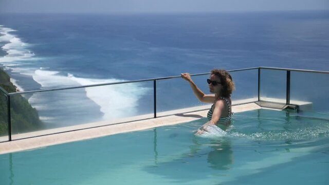 Caucasian girl splashing water in infinity edge swimming pool overlooking an ocean. Luxury holidays