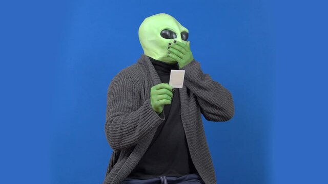 Emotional alien with green skin in human clothes looks at picture and begins to cough sitting on blue chromakey background
