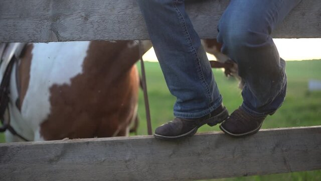 Close shot of the boots of a woman sitting on the wooden fence of a ranch with a horse behind in slow motion.
