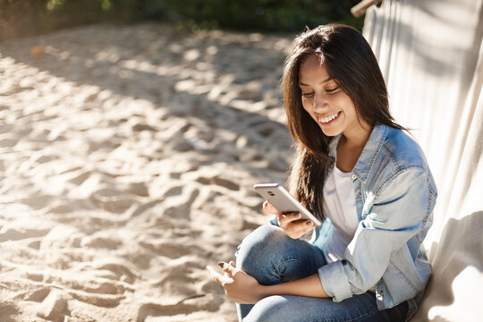 Girl writing post about beautiful sunny day on beach, edit pics from beach party as sit on hammock feet on sandy surface, smiling joyfully at smartphone screen, asking boyfriend take picture