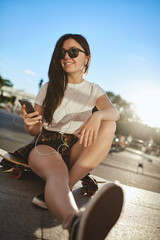 Youth, leisure and urban lifestyle concept. Vertical shot cool hipster girl with long dark hair, sunglasses, sit on wooden penny board, hold smartphone, pick playlist listen music wired earphones