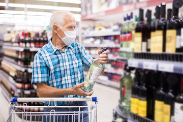 mature european man wearing mask with covid protection chooses bottle of alcohol in supermarket