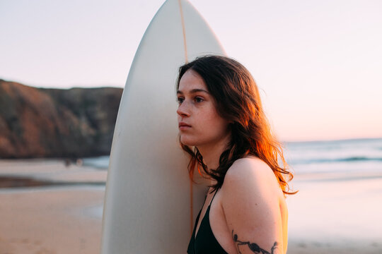Candid portrait of authentic young woman at beautiful beach, stand with surfboard and look in distance at horizon. Modern millennial nomad on vacation at surf camp or school