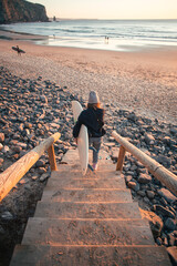 Young woman in casual clothes walk down stairs to beautiful epic beach at sunset or sunrise. Female surfer ready to start surfing lesson at dawn. Millennial lifestyle and hobby