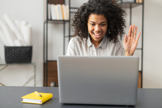 Smiling African American woman or student with Afro hairstyle sitting at the desk in the home office, studying online in the virtual classroom, saying hello to classmates, waving at the laptop screen