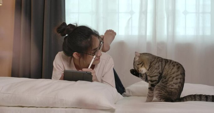 Young asian glasses woman lying on bed with cute cat. A pretty girl use tablet computer for work or study online. Lady in casual clothes at house during quarantine. Social distance new normal concept.