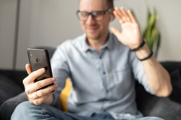 Video call concept. A smartphone in hand of a young guy, a young man in out of focus is waving hello into webcam