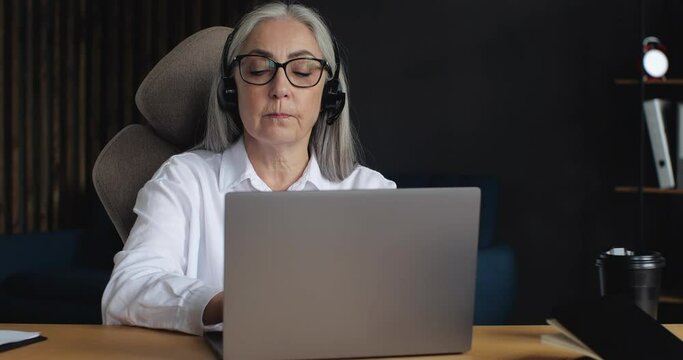 Attractive mature senior Operator in Call-Center, Technical Support is working indoor, sitting at Home Office. Happy Woman is talking speaking in Headset, using Laptop. Easy Technology.