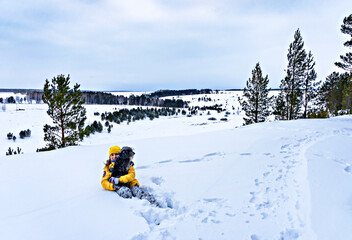 Young cheerful woman in yellow jacket sitting in snow holding fluffy gray dog on background of snowy landscape with coniferous trees. Walking, caring and traveling with pets. Winter hike, pet love