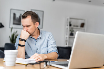 Fototapeta premium Thoughtful young man in smart casual orking with a laptop remotely, solving some tasks. A guy works online from home