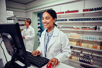 Cheerful young female pharmacist wearing labcoat working on computer in drugstore - obrazy, fototapety, plakaty