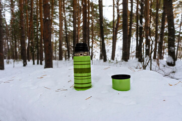 green striped thermos with hot drink standing in the snow on table in the forest. Tea drinking in nature on a winter hike, rest