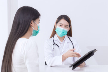 Asian woman doctor give counsel to her woman patient which doctor shows information on tablet while both wear medical face mask in hospital. Coronavirus protection concept.