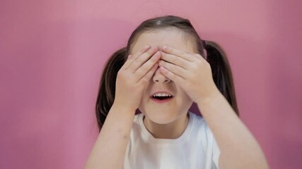 Little dark haired girl with two ponytails in white T-shirt plays hide and seek shows different emotions, makes peekaboo covering eyes with hands. Pink background. Concept of happy childhood lifestyle