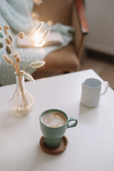 Lifestyle composition with cup of coffee, candle and a vase with flowers on a coffee table. Cozy home interior decoration concept