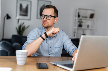 Fototapeta na wymiar An intelligent young man in glasses and casual shirt sits at the desk with a laptop and looks away lost in thoughts. A freelancer is thinking about solving business tasks