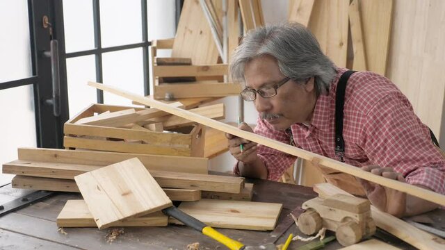 Side view, close up portrait of satisfied dedicated Asian senior male carpenter wears lumberjack shirt and apron, looking and checking the plywood slat, smiling. Retirement and business concept