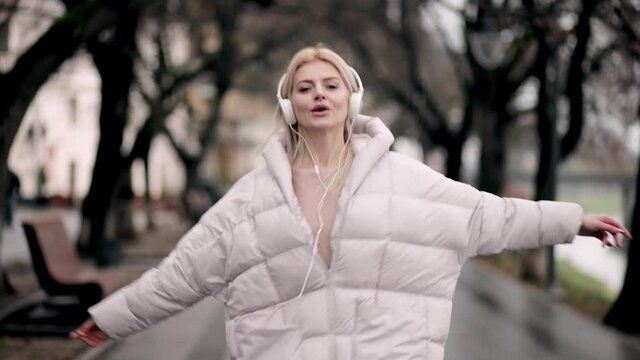Slow motion of happy young woman in headphones dancing outdoors n the city park having fun alone. Joyful attractive blonde carefree woman listening to music with smartphone