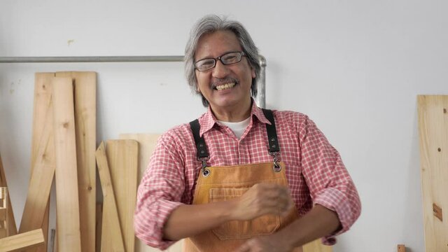 Portrait of a happy senior Asian man wearing a lumberjack shirt and apron, smiling, crossed arms, showing thumbs up, in the wood workshop. Front view, indoors, medium shot, carpenter and DIY concept