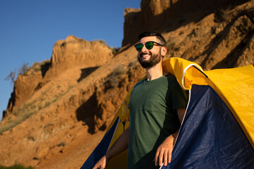 Happy young brunette bearded man tourist in sunglasses peeping out of touristic tent, smiling looking into distance, enjoying vacation adventure in camping on sandy mountains and blue sky background