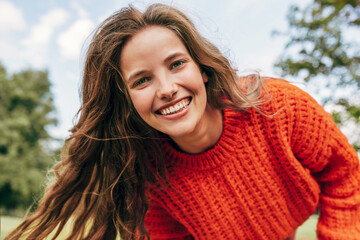 Candid portrait of a cheerful young woman wearing an orange sweater spending time in the park. The...
