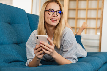 Smiling blonde woman using mobile phone while lying on couch