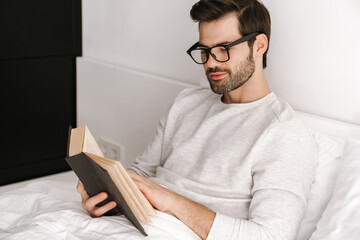 Focused young man in eyeglasses reading book while resting in bed