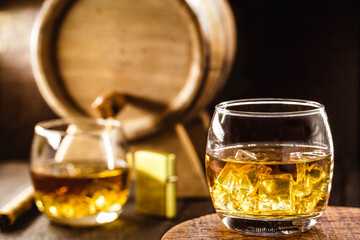 whiskey glass with ice, spot focus, bottle, lighter and barrel on blurred background. Distilled...