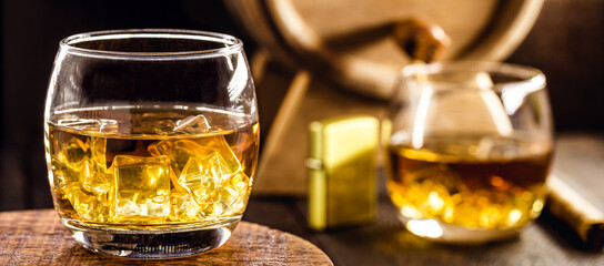 whiskey glass with ice, spot focus, bottle, lighter and barrel on blurred background. Distilled...