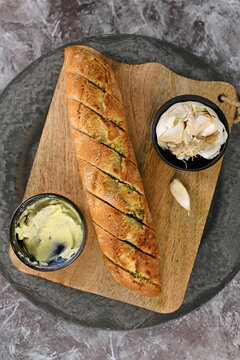 Top view of fresh Baguette bread baked and stuffed with herb butter next to bowls with herb butter and garlic on wooden cutting board