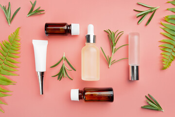 Glass and plastic cosmetic bottles on pink background with fern and rosemary leaves