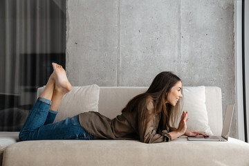 Obraz premium Cheerful brunette girl waving hand and using laptop while lying on sofa