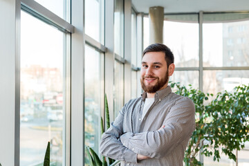 Obraz na płótnie Canvas Smiling caucasian man standing near full length window in office alone. Successful bearded young smart man crossed arms. Handsome businessman in casual shirt. Freelancer in coworking space with plants