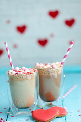Two glasses of hot coffee or cappuccino with marshmallows and heart-shaped cookies on a blue background.
