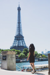 A girl in a black dress with long hair walking towards the river cruise Seine river and Eiffel Tower in Paris on a sunny day.
