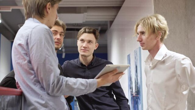Interested young Caucasian men listening to colleague explaining project using tablet. Four confident male colleagues standing in office corridor talking. Business concept.