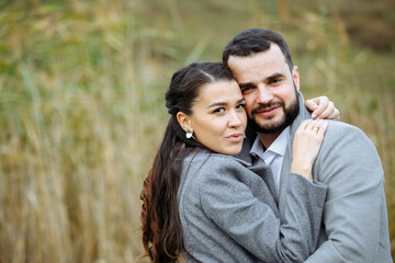Stylish couple in gray coats gently hugging on the background of reeds. Happy sensual wedding couple embracing. Romantic moments of newlyweds.