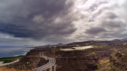 Aerial view of the mountains of the island of Gran Canaria and the coast next to the sea with clouds and fog in the background. Spain
