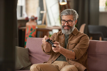 Mature bearded gamer looking exciting playing video game on sofa in the room