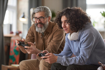 Fototapeta premium Bearded man and guy using joysticks for playing in video game while sitting on sofa at home