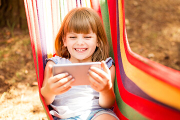 Smiling child girl recording lifestyle blog talking to camera of mobile phone outdoor, sitting on hammock. Happy young influencer filming vlog for her channel. Blogging Concept. 