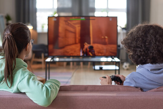 Rear View Of Two Teenagers Sitting On Sofa In Front Of Big Screen Of TV And Playing Video Game At Home